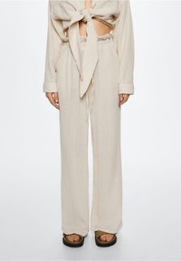 Ensemble léger et texturé de couleur crème, comprenant un crop top noué et un pantalon plissé ample, complété par des sandales plates.