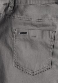 Poche arrière en tissu gris de forme rectangulaire, dotée d'un bouton en métal et d'une petite étiquette de marque au-dessus de la couture de la poche.