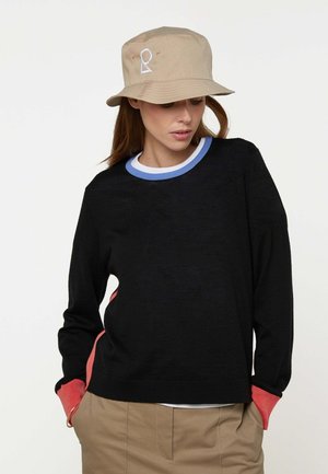 Vrouw met beige vissershoed met wit logo, zwarte trui met blauwe en witte halslijn, koraalkleurige boorden en beige broek, handen in de zakken.