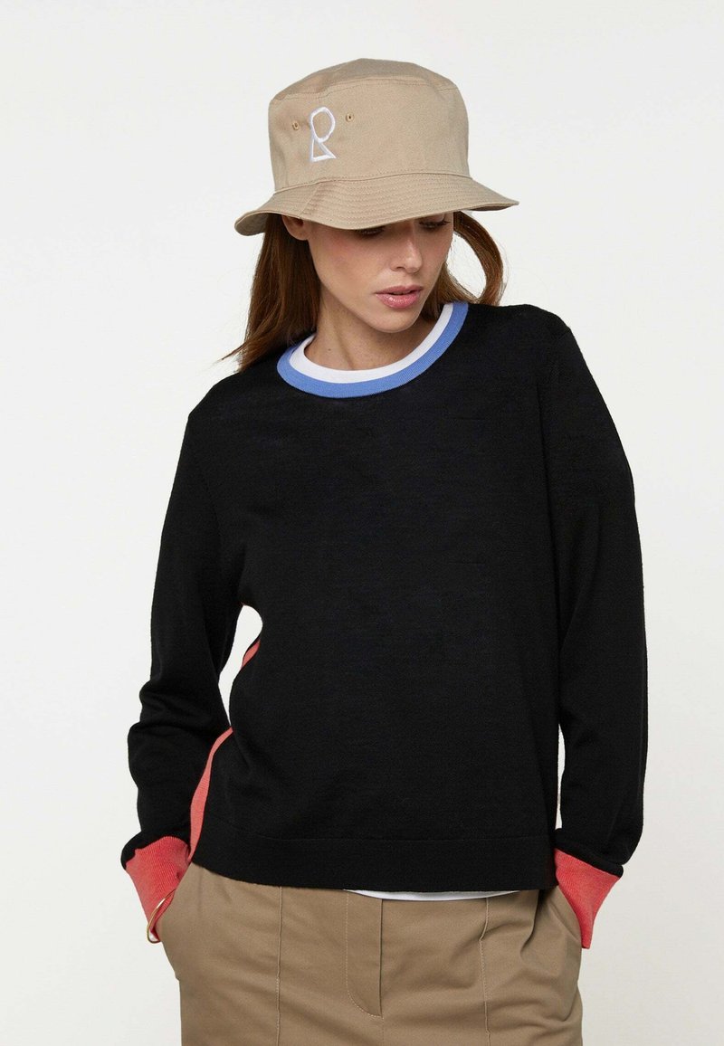 Frau trägt einen beigen Anglerhut mit weißem Logo, einen schwarzen Pullover mit blau-weißem Kragen, korallfarbene Bündchen und beige Hose, Hände in den Taschen.