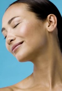 Gros plan du visage d'une femme avec les yeux fermés, peau lisse, léger blush et couleur naturelle des lèvres, sur fond bleu clair uniforme.