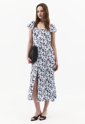 Jonge vrouw draagt een midi-jurk met witte en marineblauwe bloemenprint, pofmouwen en een voorste split, houdt een zwarte clutch vast en draagt zwarte slides.