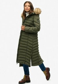 Lang olijfgroene donsjas met een chevron-quiltpatroon, bontomzoomde capuchon en ritsclosure. Combinatie met blauwe jeans en bruine veterschoenen.