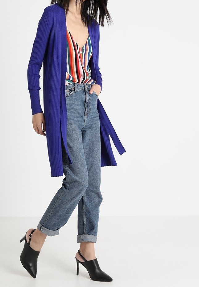 Femme portant un long cardigan bleu, un chemisier rayé multicolore, un jean retroussé et des chaussures à talons slingback noires, debout sur fond blanc.