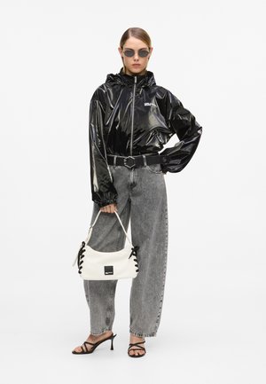 Žena u crnoj sjajnoj jakni s kapuljačom, sivim trapericama visokog struka, crnim sandalama na petu, sunčanim naočalama i drži bijelu torbu s crnim vezicama.