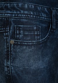 Dark blue denim jeans close-up focusing on the front small coin pocket and surrounding stitching details.