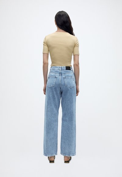 Γυναίκα με μακριά σκούρα μαλλιά που φοράει μπεζ κοντομάνικη μπλούζα, ανοιχτό μπλε τζιν με φαρδύ παντελόνι ψηλόμεσο και μαύρα πέδιλα με ανοικτά δάχτυλα, στέκεται με την πλάτη προς τα εμπρός.