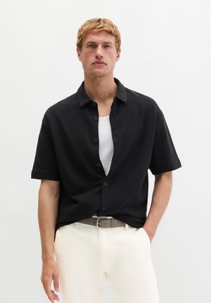 Junger Mann mit kurzen blonden Haaren trägt ein schwarzes kurzärmliges Hemd mit Knöpfen über einem weißen Top und weißen Hosen mit einem beigen Gürtel.