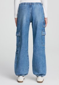 Luźne niebieskie dżinsowe spodnie cargo z wysokim stanem, elastycznym pasem i kieszeniami na kolanach. Posiadają marszczenia na kostkach oraz białe trampki.