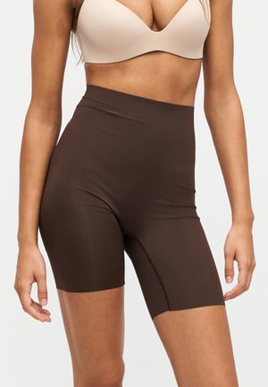 Femme portant un cuissard de cyclisme marron foncé taille haute et un soutien-gorge beige, debout devant un fond clair uni.
