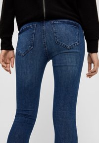 Jeans de mezclilla azul oscuro con un ajuste slim, que cuentan con dos bolsillos traseros y una ligera textura. Llevados con una blusa negra, con un detalle de cremallera visible.