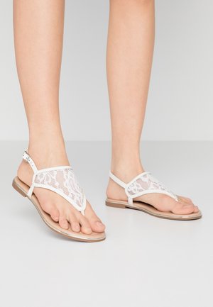 Sandales blanches en dentelle avec un design en V, dotées d'une semelle plate et d'une sangle à la cheville. Le détail en dentelle texturée ajoute un intérêt visuel.