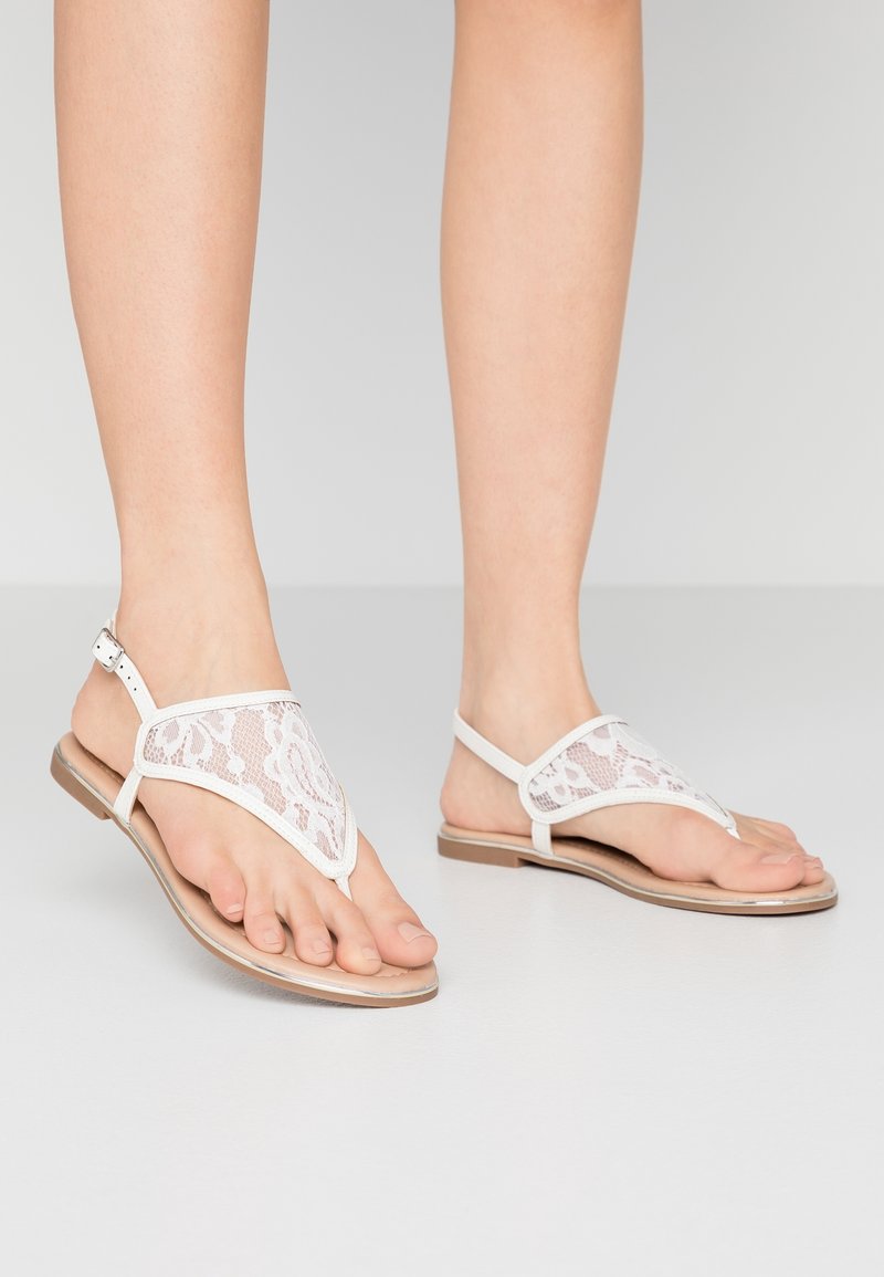 Valkoiset pits sandaalit V-muotoisella muotoilulla, joissa on litteä pohja ja nilkkahihna. Texturoitu pitsiyksitys lisää visuaalista kiinnostavuutta.