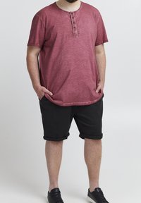 Homme debout, les mains dans les poches d'un short noir, portant une chemise rouge à manches courtes boutonnée et des baskets noires, sur un fond blanc.