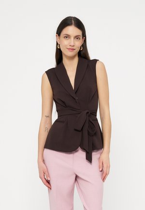 Žena s dlhými tmavými vlasmi, ktorá má na sebe tmavohnedý bezrukávový zavinovací blejzer zviazaný v páse a svetloružové nohavice, stojaca na bielom pozadí.