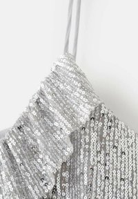 Svetlikasta bluzica s srebrnimi paščki in nabranim dizajnom, ki prikazuje sijočo, teksturirano površino na nevtralni podlagi.