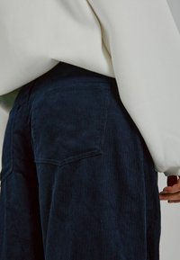 Marineblaue Cordhosen mit vertikalen Rillen, ausgestattet mit einer Gesäßtasche, kombiniert mit einem cremefarbenen Pullover, der über den Taillenbund fällt.