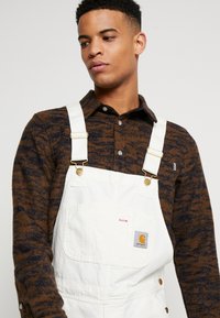 Salopette blanche avec des bretelles ajustables, des accessoires en or et une poche frontale. Associée à une chemise à boutons à motifs marron et bleu marine.