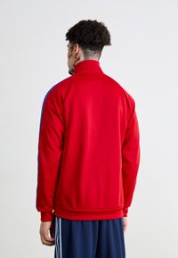 Giacca da corsa rossa con colletto alto, caratterizzata da strisce laterali blu. Realizzata in un tessuto liscio con un design aderente. Orlo e polsini cuciti.