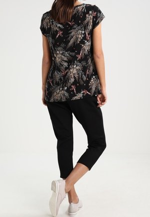 Kobieta nosząca czarną, kwiecistą bluzkę z krótkim rękawem, czarne spodnie 3/4 i jasnoróżowe trampki, stojąca na tle jednolitego, jasnego tła.