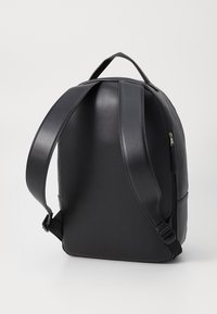 Svart läderryggsäck med slät yta, något böjd form, justerbara remmar och en ficka med dragkedja på sidan.