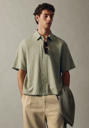 Jeune homme portant une chemise verte à carreaux à manches courtes, tenant un pull gris, avec des lunettes de soleil accrochées au col de la chemise.