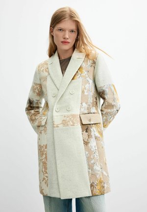 Junge Frau mit langen blonden Haaren, die einen beigen und cremefarbenen Patchwork-Blumen zweireihigen Mantel über Jeans trägt, steht vor einem schlichten Hintergrund.
