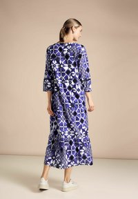 Lång, flödande klänning i lila och vit geometrisk mönstring. Trekvartsärmar med en design av flera lager i kjolen. Texturerad tyg, avslappnad stil.