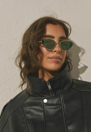 Žena s vlnitými hnedými vlasmi, ktorá má na sebe zelené slnečné okuliare v tvare mačacích očí a čiernu koženú bundu s vysokým golierom, stojaca pred textúrovanou béžovou stenou.