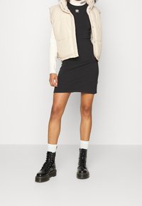 Beige pufferväst över en svart figurnära klänning, kombinerad med svarta snörstövlar och vita ankelsockor. Vesten har en huva och tryckknappar.