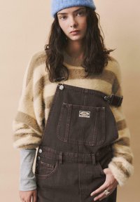 Braune Denim-Latzhose mit einer Fronttasche, silberner Hardware und einem Etikett. Kombiniert mit einem beigen gestreiften, flauschigen Pullover und einer hellblauen Mütze.