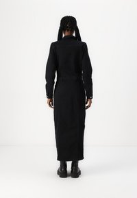 Robe en denim noir avec une coupe longue et ajustée et des manches à boutons. Présente une taille cintrée et une coupe droite jusqu'aux chevilles. Bottes noires épaisses.