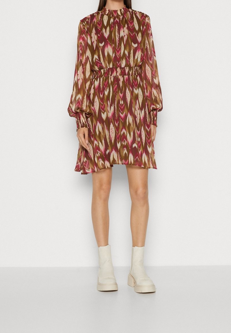 Femme portant une robe à manches longues, au-dessus du genou, avec un motif abstrait rouge, marron et beige, associée à des bottines blanches.