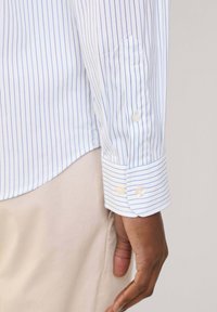 Camicia bianca a maniche lunghe con righe verticali blu, caratterizzata da polsini con bottoni e una texture liscia. Il tessuto è leggero e informale.