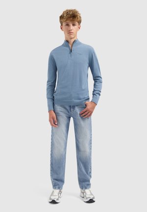 Blaues Langarm-Pullover mit einem halben Reißverschluss am Kragen, kombiniert mit hellblauen, ausgewaschenen Jeans und silbernen Sneakers. Schlichtes Design, weicher Stoff.