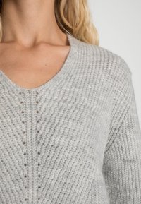 Pull en tricot gris clair avec un col en V, texture côtelée et détails de point ouvert subtils sur le devant. Tissu doux et confortable.