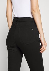 Primer plano de una persona que lleva vaqueros negros de cintura alta y una camiseta sin mangas blanca con texturizado, mostrando el detalle del bolsillo trasero y la mano descansando al costado.