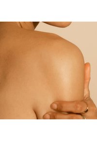 Peau lisse et bronzée d'une épaule avec une main reposant dessus. La peau présente une texture douce et un éclat subtil, le tout sur un fond atténué.