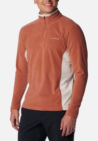 Mann, der einen rostfarbenen Fleece-Pullover mit cremefarbenen Seitenpanelen und einem halben Reißverschluss trägt, kombiniert mit dunklen Hosen, und lächelt mit entspannten Händen.