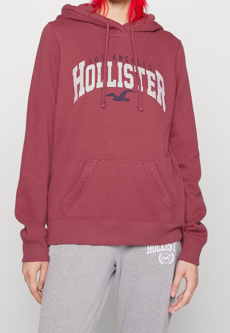 Sudadera con capucha color burdeos y un bolsillo frontal, con un logo blanco de "Hollister" y detalles en gris. Material suave, puños y dobladillo acanalados.
