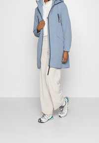 Chaqueta impermeable de color azul claro con capucha, presenta un cierre frontal y bolsillos laterales. Combinada con pantalones beige y zapatillas de colores.