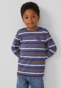 Jongentje met krullend haar draagt een gestreepte lang gedragen shirt in blauw, bordeauxrood, wit en olijfgroen, en blauwe jeans, staand tegen een effen achtergrond.
