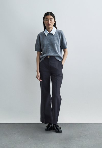 Femme portant un pull à manches courtes gris par-dessus une chemise à col rayée, un pantalon noir à jambes larges et des mocassins noirs, debout contre un mur gris.