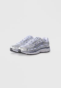 Un paio di scarpe da corsa Nike argento e bianche con suole nere e dettagli blu, esposte su uno sfondo grigio chiaro uniforme.