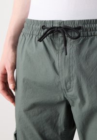 Jasnoszaro-zielone spodnie z elastycznym pasem i czarnym sznurkiem, o delikatnej prążkowanej fakturze oraz bocznych kieszeniach.