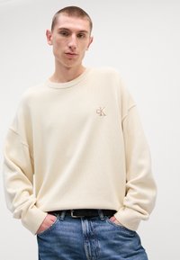 Μπεζ πουλόβερ πλεκτού τύπου με ραμμένο κολάρο και μανσέτες, διαθέτει κεντητό λογότυπο cK στο στήθος. Συνδυασμένο με μπλε τζην παντελόνι.