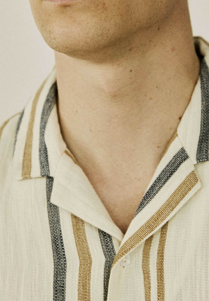 Kortærmet skjorte lavet af struktureret beige stof med lodrette striber i blå og brun. Har en spids krave og knaplukning.