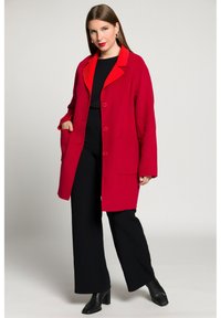 Cappotto in misto lana rosso con colletto rosso brillante, chiusura a un solo bottone, due tasche anteriori, vestibilità rilassata, abbinato a pantaloni neri a gamba larga e stivali.