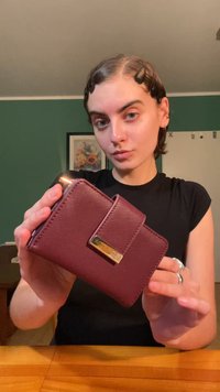 Burgundijas mākslīgās ādas maks ar teksturētu virsmu, magnētisko aizdari un zelta krāsas akcenta plāksni priekšā, ko tur cilvēks.