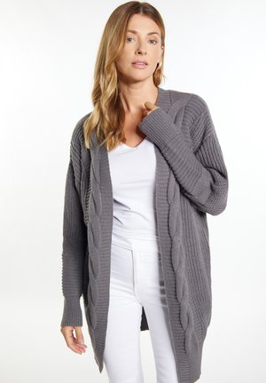 Šedý pletený cardigan s žebrovanými rukávy a otevřeným předním designem, vrstvený přes bílý top s měkkým, texturovaným povrchem.